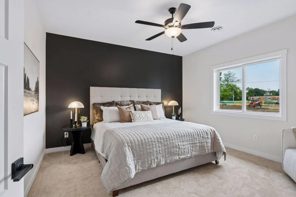 A modern bedroom featuring a black accent wall behind a light gray upholstered bed, neutral bedding, matching black nightstands with lamps, a ceiling fan, and a window showing an outdoor view.
