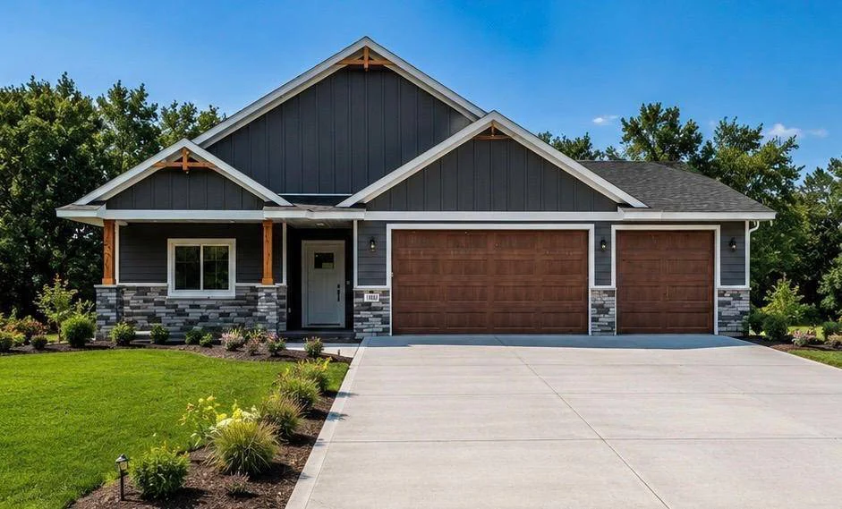 A single-story home featuring dark gray siding with white trim, a three-car garage with brown wood-look doors, stone veneer accents along the foundation, natural wood porch pillars, a wide concrete driveway, and a landscaped green lawn.