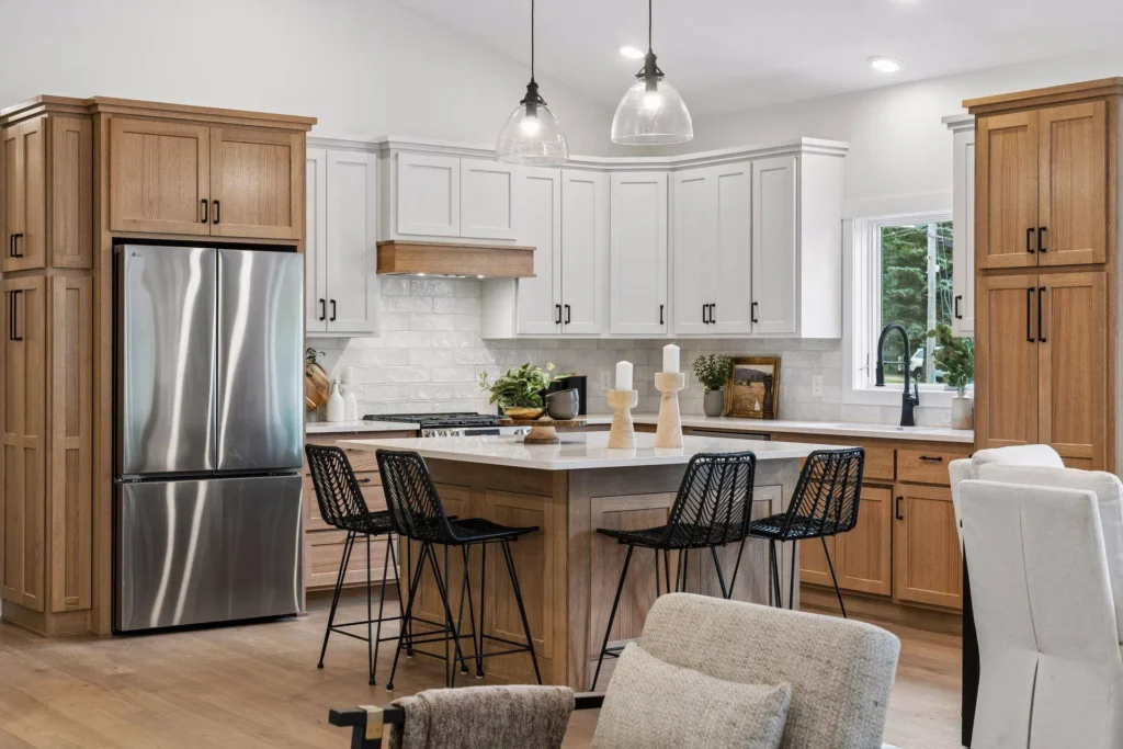A modern kitchen featuring a large wood center island with a white countertop and four black wire barstools, natural wood and white cabinetry, a stainless steel refrigerator, white subway tile backsplash, and light wood flooring.