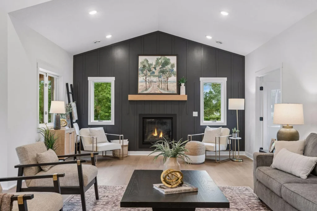 A bright living room featuring a vaulted ceiling, a dark charcoal board-and-batten accent wall with a fireplace and natural wood mantel, flanked by two white-framed windows. The space is furnished with a gray sofa, two light-colored patterned armchairs, a black square coffee table resting on a patterned area rug, and light wood flooring.
