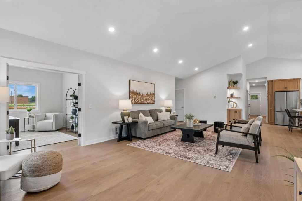 A spacious, open-concept living room featuring a vaulted ceiling with recessed lighting, light wood flooring, a gray sofa, two wood-framed accent chairs, a black coffee table resting on a patterned area rug, built-in wall shelving, and wide entryways leading into a bright adjacent sunroom and the kitchen.