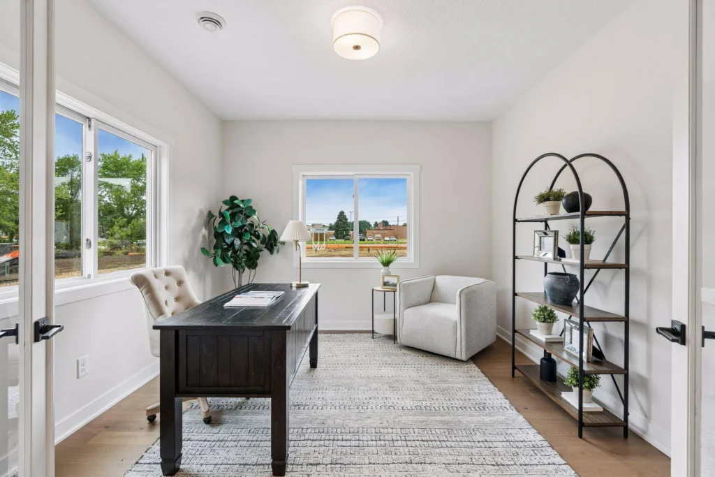 A bright home office featuring light wood flooring, white walls, and a dark wood desk resting on a textured area rug. The room includes a beige tufted desk chair, a light gray accent chair in the corner, a tall, arched black metal and wood open bookshelf displaying decor and plants, and two white-framed windows letting in natural light.