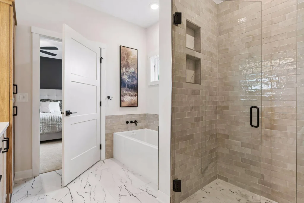 A bright en suite bathroom featuring a large glass-enclosed shower with light textured subway tile and two built-in niches, a white bathtub with black wall-mounted fixtures, large white marble-patterned floor tiles, natural wood cabinetry, and an open white door leading to the adjacent bedroom.