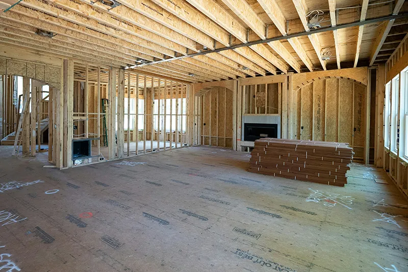 The interior framing of a new residential home under construction, showing exposed wooden wall studs, ceiling joists, and a large window opening.