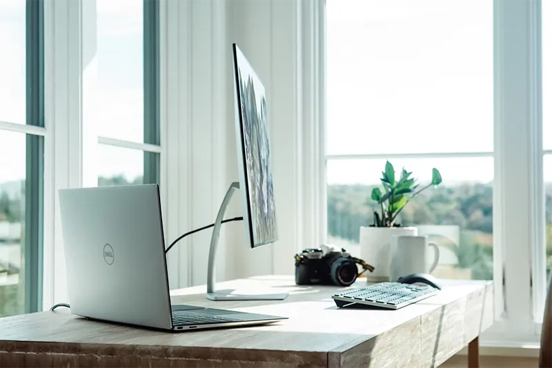 A bright, modern home office workspace featuring a white desk, a desktop computer monitor, a small potted plant, and framed artwork next to a window with natural light.