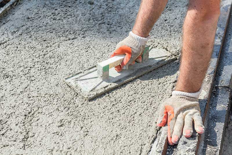 A construction worker wearing work boots and gloves kneeling to smooth wet concrete along the edge of a new home's slab foundation.